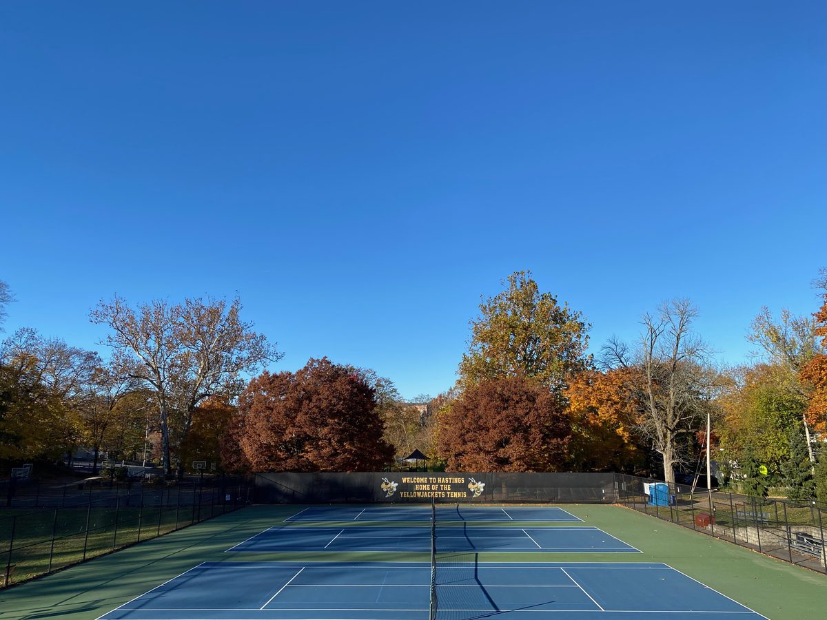 Blue tennis courts with a Welcome to Hastings banner, backed by red, orange and green trees under a clear sky